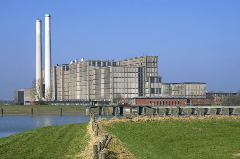 Een grote, grijze elektriciteitscentrale met twee schoorstenen, te zien aan de overkant van een sloot.