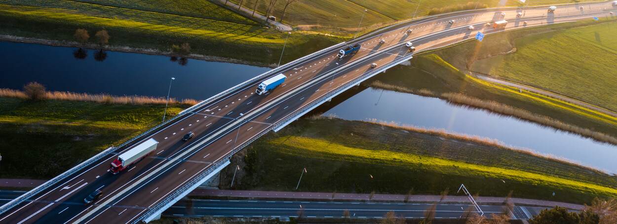 Beeld van een snelweg over een water en groen, met vrachtwagens en auto's.