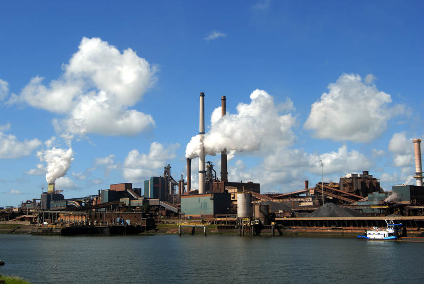 Foto van Tata steel aan de overkant van het water, uit sommige schoorstenen komt rook.