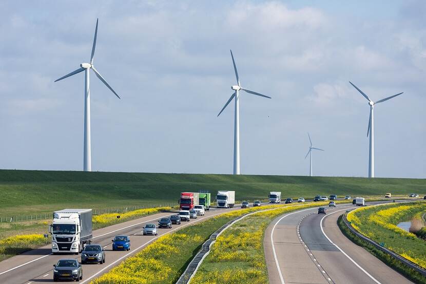 Twee autowegen lopen langs elkaar door het groen, met auto’s en vrachtwagens op de wegen. Op de achtergrond zijn een aantal windmolens zichtbaar.
