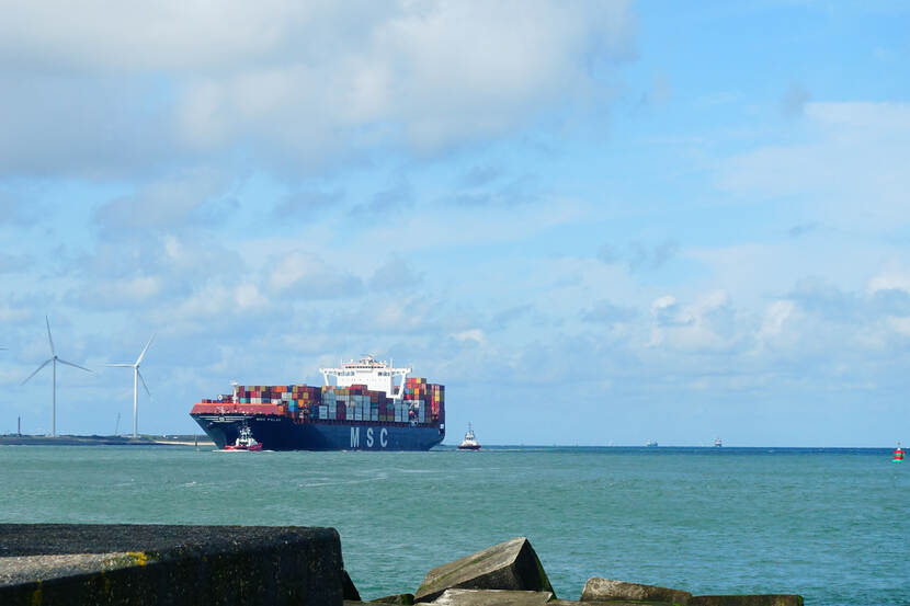 Een volgeladen containership op zee, vlakbij de kust. Op de achtergrond zijn een paar windmolens zichtbaar.