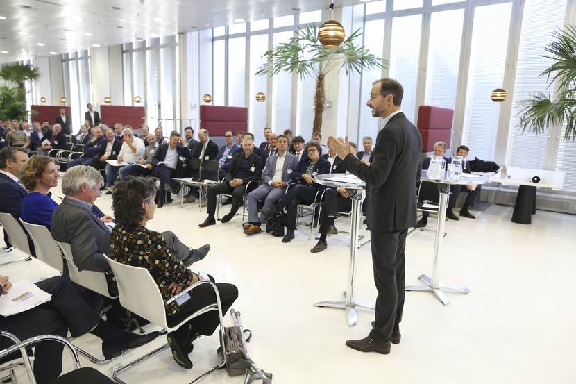 Foto van de netwerkbijeenkomst. Een aantal mensen zitten aan beide kanten en luisteren naar een collega die hen toespreekt.